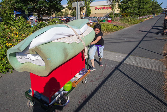 A man pushes a cart with a number of items down a sidewalk.