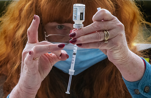 Barbara Hoffman, a mask-wearing middle-aged woman with firey red hair and red fingernails, a community health program manager and registered nurse, taps bubbles out of a syringe filled
          with COVID-19 vaccine as teachers and others from the North Kitsap School District line up in their cars in the parking garage at the
          Suquamish Clearwater Casino Resort in Suquamish, waiting to be vaccinated.