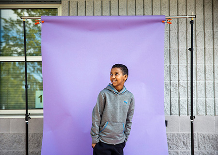 Fifth-grade student Jabir Ahmed is photographed at Kent Elementary School on Friday, May 21, 2021. He is smiling and gazing upward out of frame in front of a purple backsheet palced against the school.